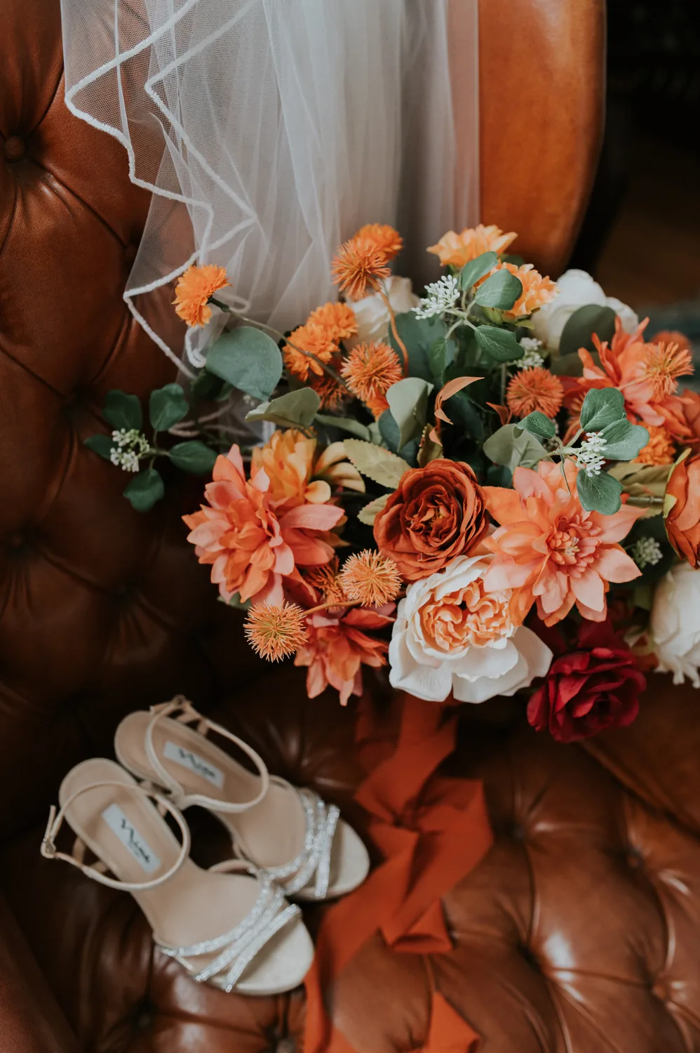 Orange, ivory and burgundy bouquet with eucalyptus-style greenery, styled with bridal accessories.